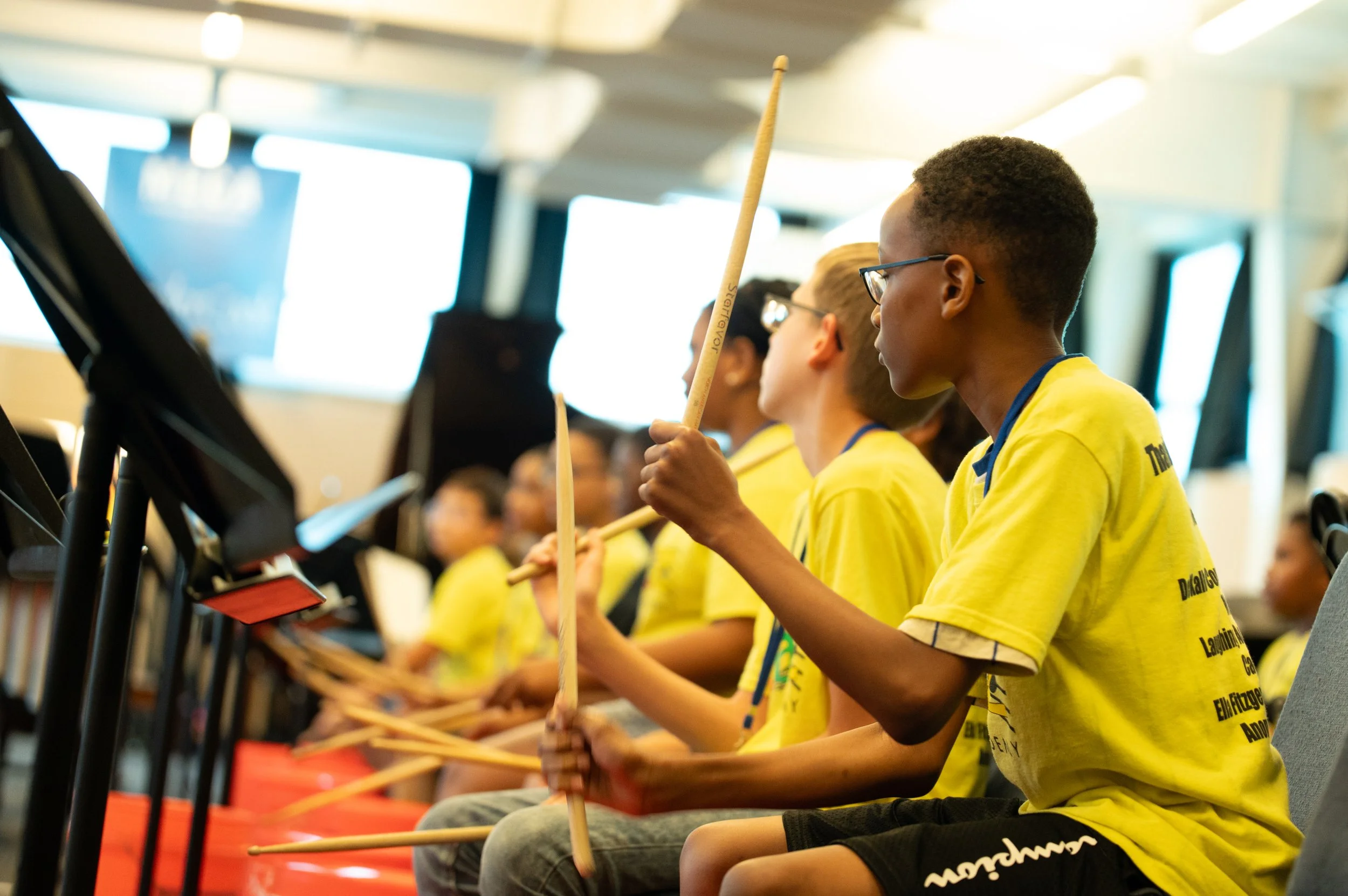 Students in yellow Jazz Academy shirts playing along on drumsticks in a rehearsal room, music stands in front of them